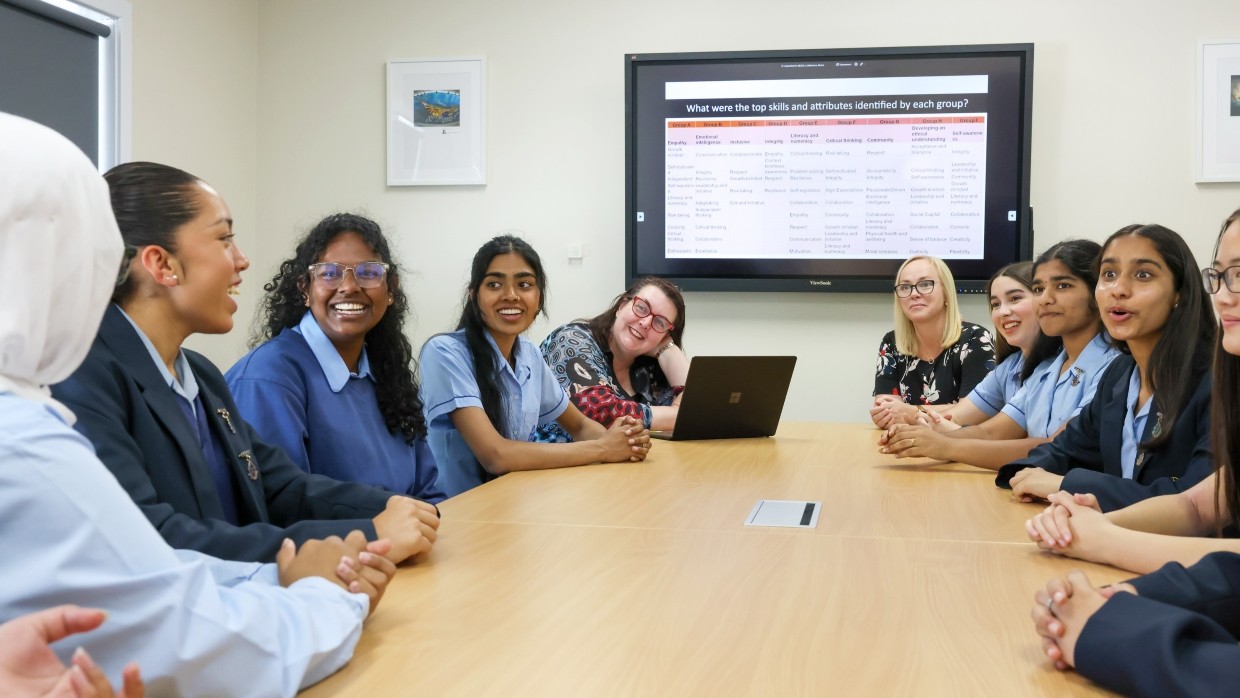 Group of 8 female high school students at desk talking with school executive.