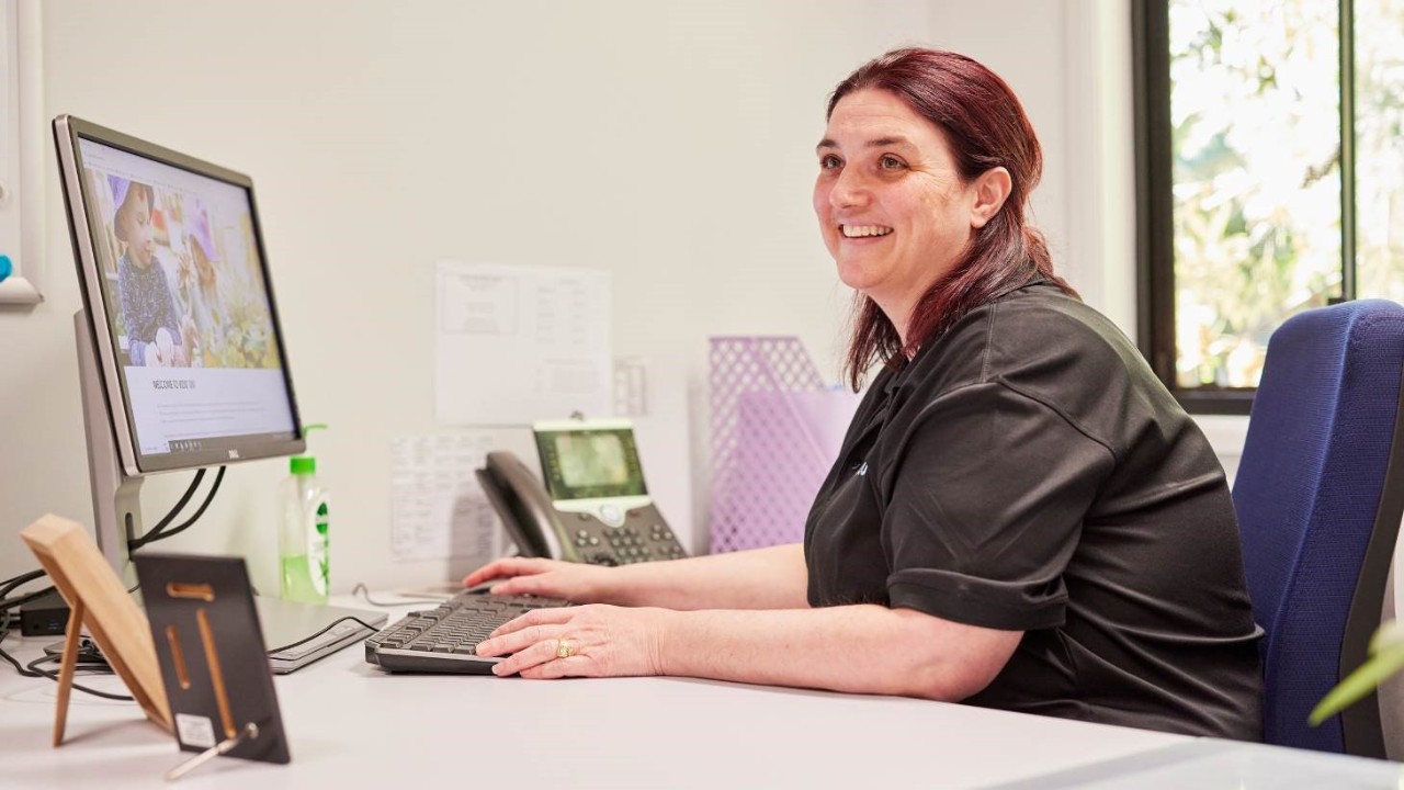 A female educator looking up and smiling while sitting at a desk in front of a computer and keyboard.