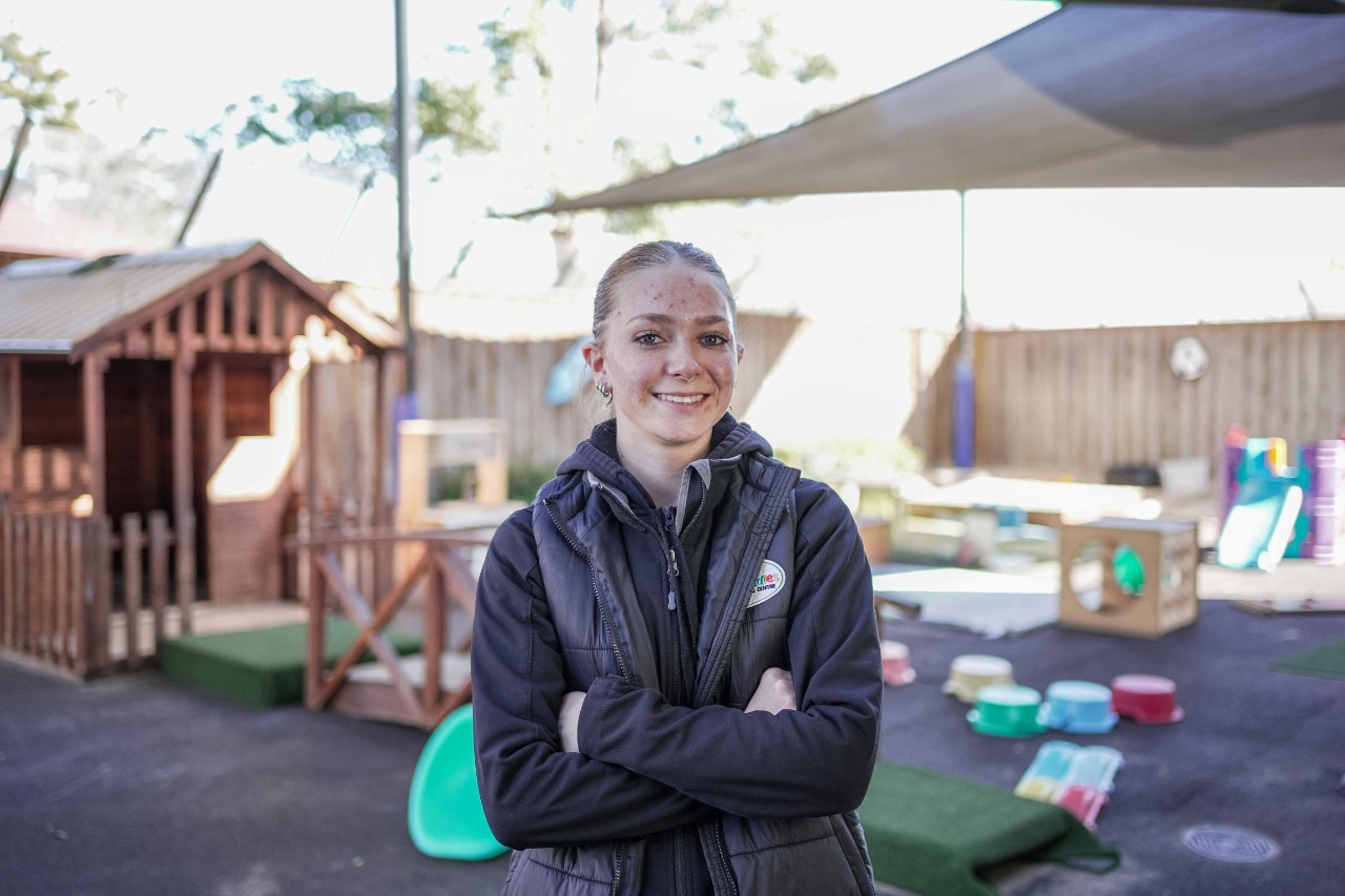 A young woman stands in front of an outdoor playground smiling. She is wearing a long black jumper and short sleeve puffer jacket.