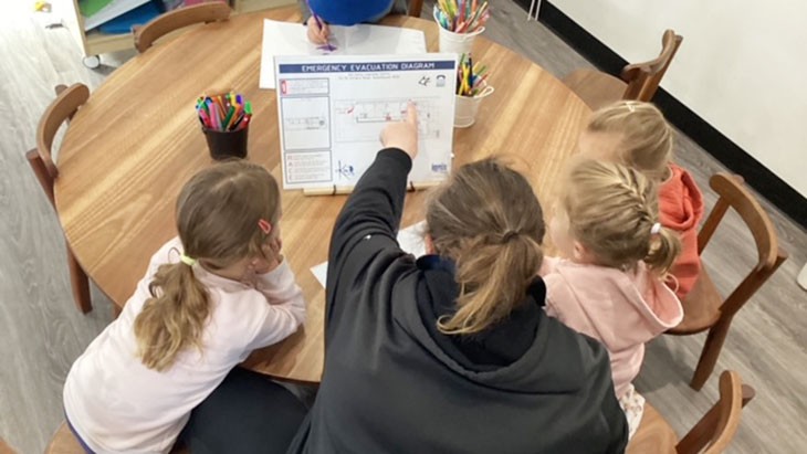 An educator wearing a black hoodie sits at a round wooden table next to 3 small children. She is pointing at an emergency evacuation diagram, which is propped up on the table in front of her, while the children look on.