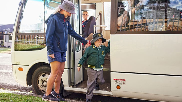 A female educator stands on a curb, watching as a child wearing a school uniform disembarks a minibus.