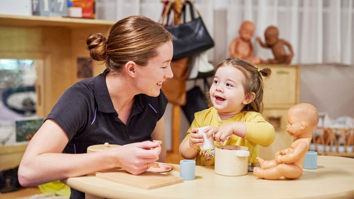 A young child wearing a bright yellow top smiles up at a female educator who is sitting beside her. The child and educator are engaged in imaginative play using toy kitchenware. A baby doll sits on the table in front of them.
