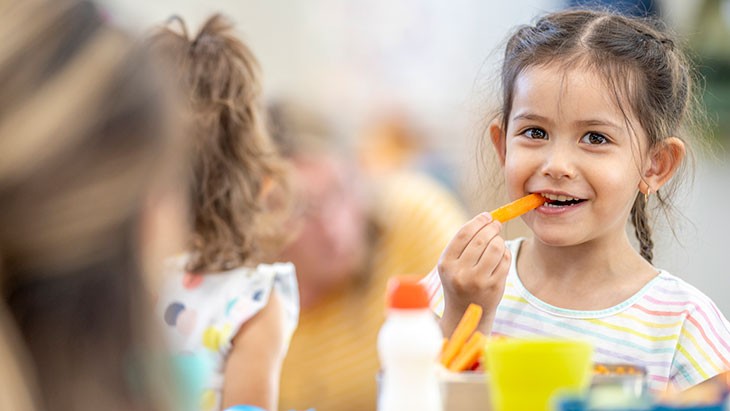 A young female child smiles while biting on a carrot. A yellow cup water bottle and container of carrot sticks sits on a table in front of her.