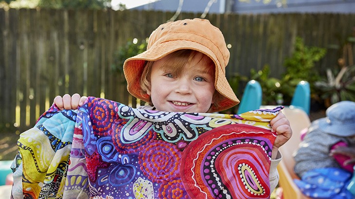 A young child wearing an orange hat smiles as they hold a piece of fabric featuring brightly coloured Aboriginal artwork.