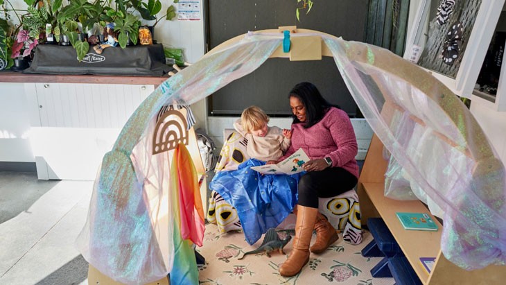 A family day care educator reads to a child in a cosy wooden reading nook.