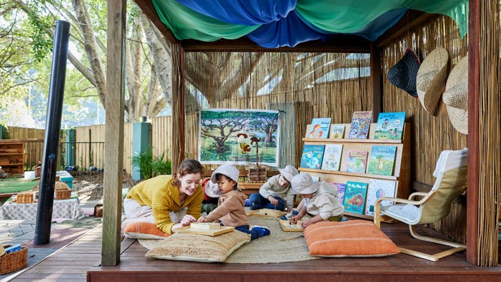 An educator sits next to a young child, who is playing with wooden toys sitting on a large cushion. They are sitting in a covered outdoor reading and play area, with an armchair, book display shelves and a round woven rug.