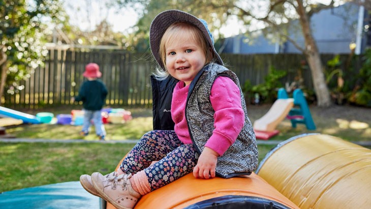 A young child with short blonde hair sits on padded play equipment in a green, leafy backyard. The child is wearing floral tights, a bright pink jumper, a dark blue patterned vest and a bucket hat. In the background, another child walks toward a path of colourful stepping stones. A small yellow and green slide is also set up in the backyard.