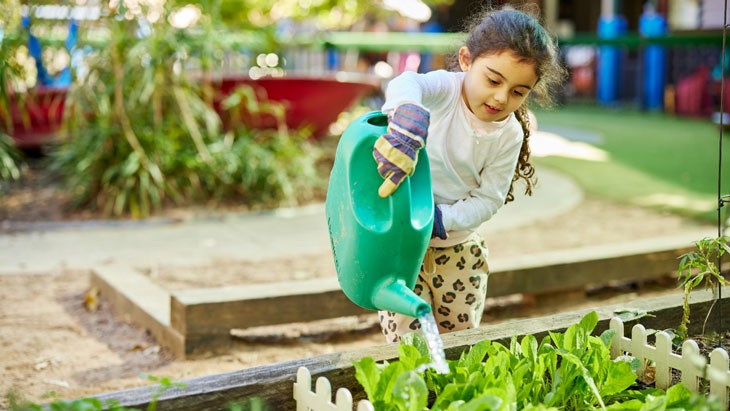 Young girl in a white shirt and leopard printed pants waters a veggie patch with a green watering jug in an outdoor space of the education service.