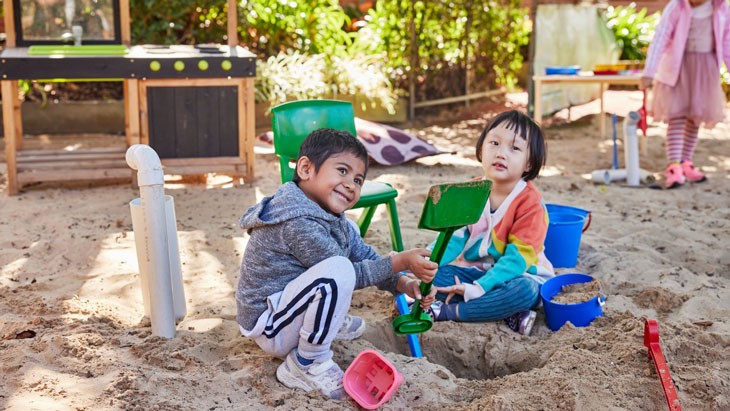 An outside of school hours care educator sits in between 4 school-aged children on an outdoor bench seat.