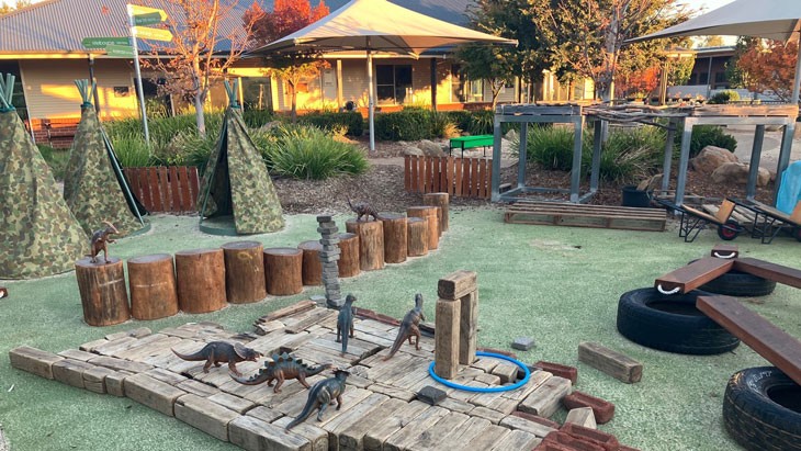 An outdoor space with mod grass, play tents, plastic dinosaur toys and oversized wooden play toys.