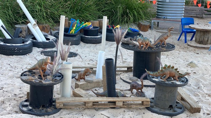 A sandpit area with a variety of large loose parts for play, including spools, wheel tyres, wooden planks, tubes and plastic dinosaur toys.