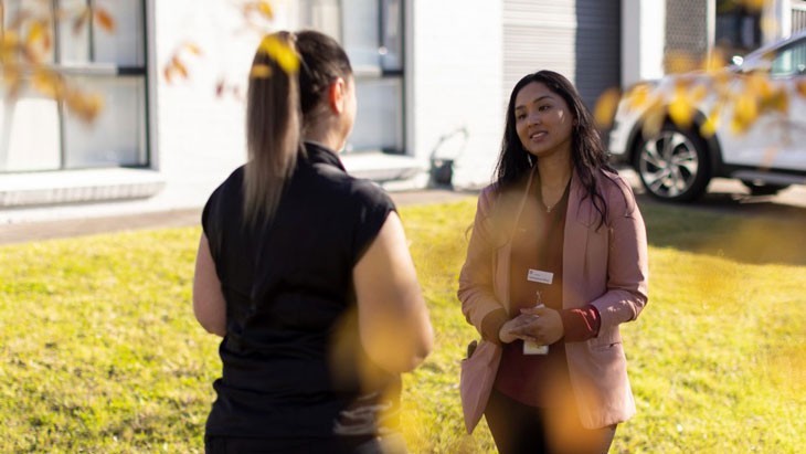 A female authorised officer speaks with an educator outdoors. The authorised officer has long dark hair and is wearing a light pink blazer, with a red long-sleeve top and black trousers. She has a lanyard with a name badge around her neck. The educator wears a black short-sleeved top and black trousers. Her long hair is in a ponytail.