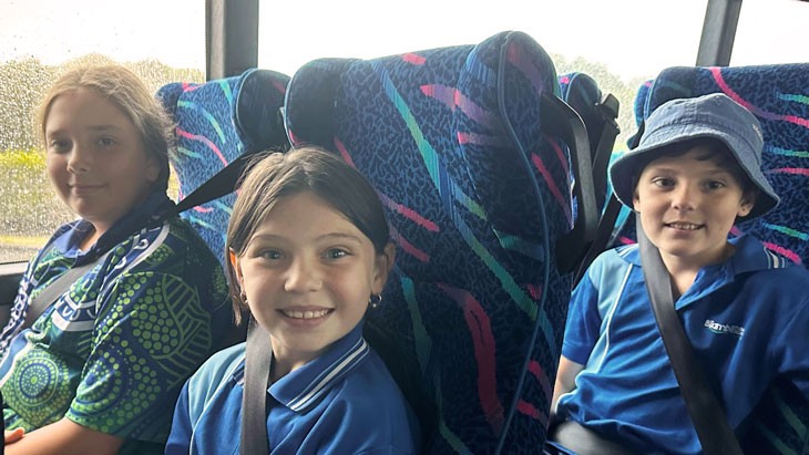 Three smiling school-aged children are seated on a bus. They each wear lap sash seat belts.