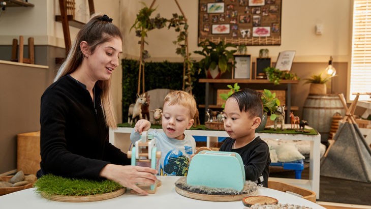 An educator wearing a long-sleeved black top sits next to 2 small children at small round table. Her long blonde-brown hair is in a high ponytail. The 2 children, who both have short hair and wear long-sleeved shirts, play with wooden toys at the table. The room is decorated in neutral colours and features several plants, vines and other greenery.