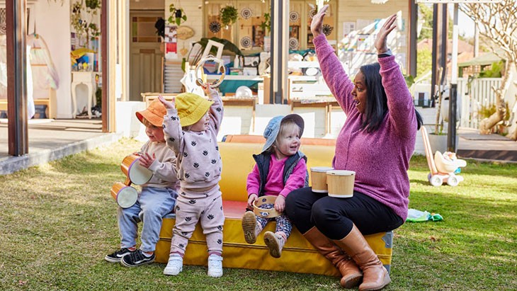 A female family day care educator sits on the right edge of a yellow and red cushioned mat in a backyard. She is engaging with 3 young children who are also positioned along the mat, while holding her hands in the air. A set of small bongo drums sits on her lap. The small children are holding tambourines or bongo drums.