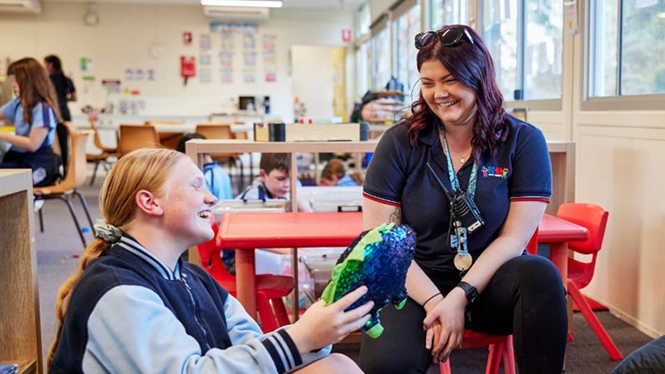 An outside school hours care educator and school-aged child wearing a light and dark blue school uniform sit together chatting in a classroom. They are both smiling. The child holds a round blue and green sequined toy in her hand. Other children are engaging in activities in the background.