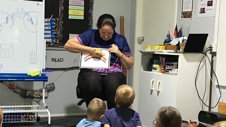 An educator sits on a chair in front of a group of preschoolers drawing on a white piece of paper with a yellow marker pen. A red, orange, yellow and purple rainbow covers most of the page, which has a thick black border.