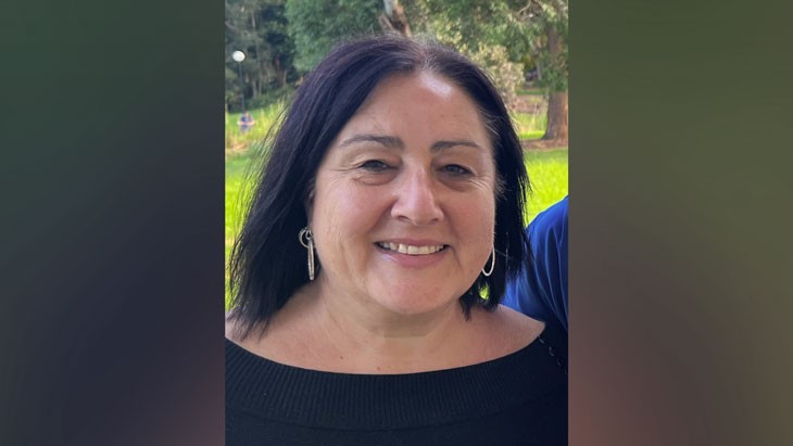 A headshot of a woman with dark shoulder length hair, wearing a black top and silver earrings. She is standing outdoors in a green, leafy area.