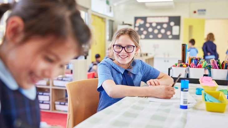 A happy school student wearing glasses and a blue tunic sits at a table drawing. Another student, who is also smiling, is visible in the foreground.