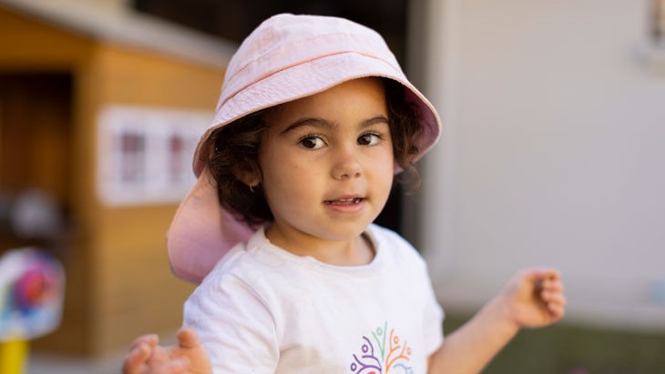 A young child standing in an outdoor play area with a pink hat.