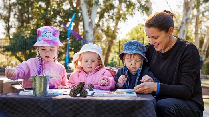 Three children and an educator sit at an outdoor table painting.