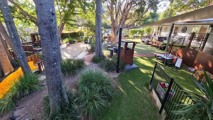 A large, natural outdoor learning area, with play equipment, a large sandpit and multiple seating areas for children. Tall trees and other leafy plants are scattered across space, providing shade over the area.