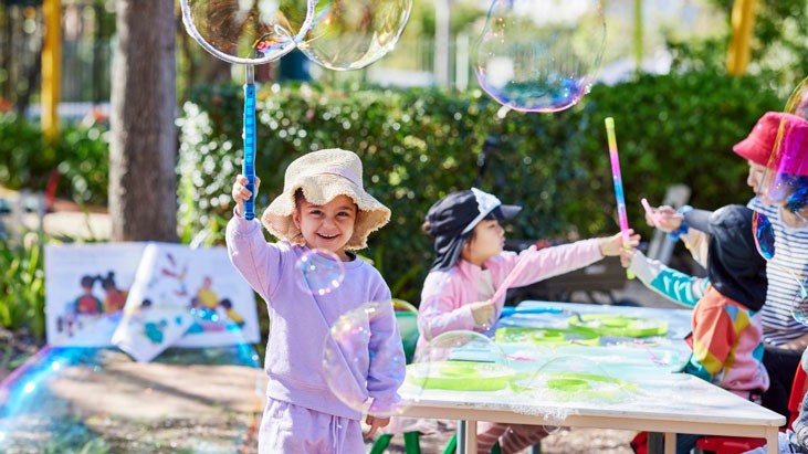 A young child wearing a light purple tracksuit and wide brimmed hat stands outdoors playing with a bubble wand. Several large bubbles float in the air around her, while two children and an educator seated at a table in the background are also engaging in bubble play.