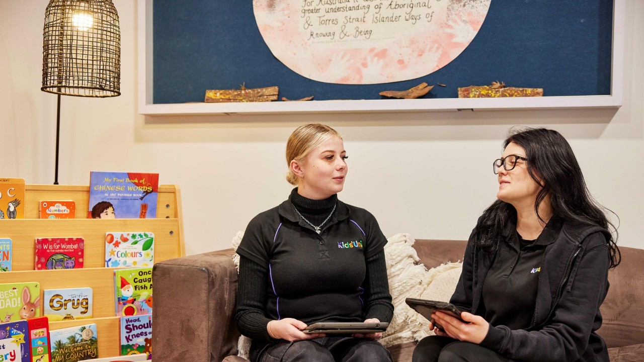 Two early childhood educators sitting on a lounge talking while holding tablets