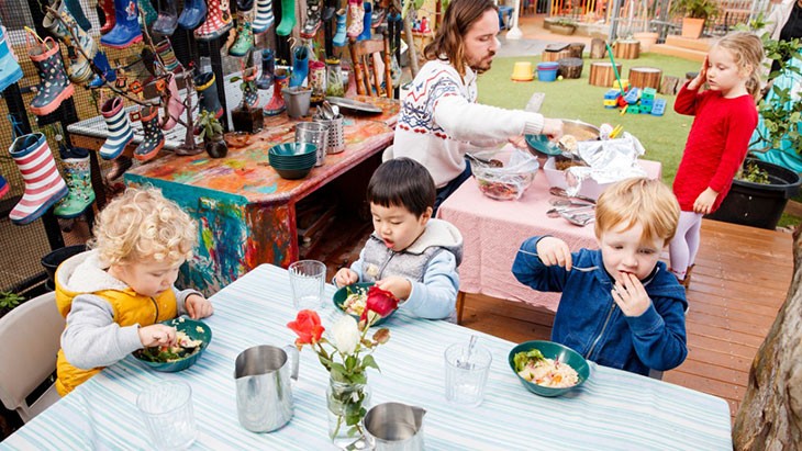 Three Children sitting at a table eating with their teacher serving food to another child who is standing up.