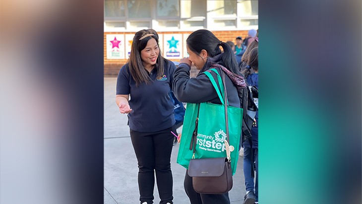 A person standing, smiling and talking to another person.