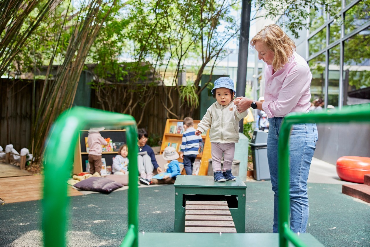 Educator helping a child across a bridge on outside paly equipment. There is an educator with 4 children in the background reading books.