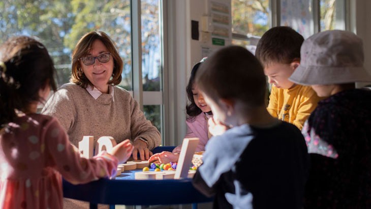 Female educator sitting at a circular table smiling at five children also sitting at the table facing her with their backs to the camera.