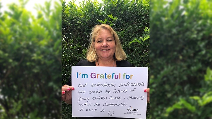 Woman with shoulder length blond hair holding a landscape poster with a heading I am grateful for. Handwritten text under the heading reads our enthusiastic professionals who enrich the futures of young children, families and students within the communities we work in. A smiley face has been drawn at the bottom of the poster.