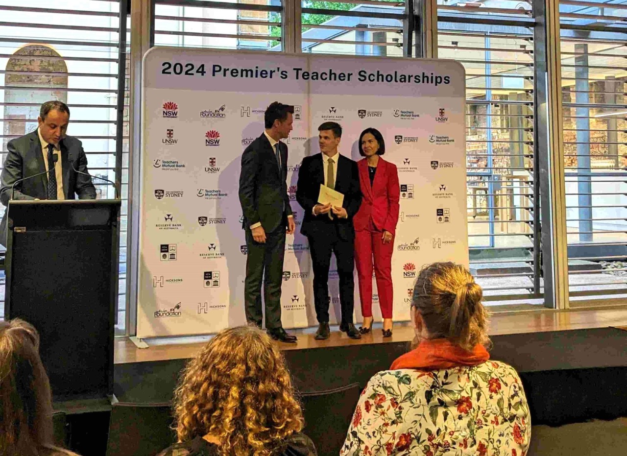 Alex Sutherland is standing on stage with Premier Chris Minns and Deputy Premier Prue Car to accept his scholarship award.