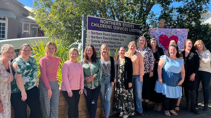 15 Northern Rivers Family Day Care staff stand in line out the front of their office sign. One woman on the right towards the back holds a flag with a large red heart in the centre.