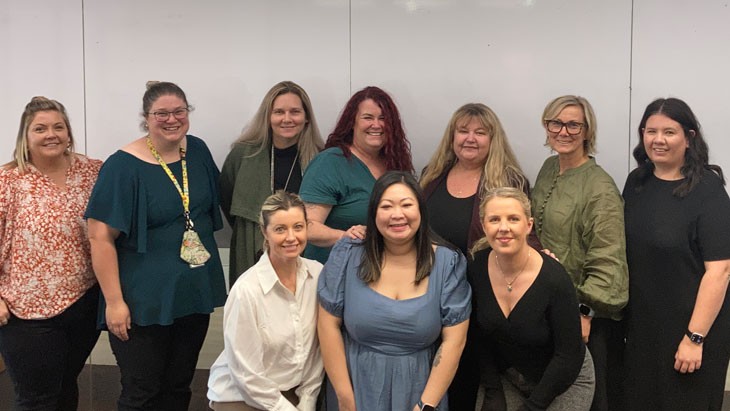 10 women of compliance team 2 stand in an office in 2 lines with 3 women kneeling at the front and 7 standing at the back.