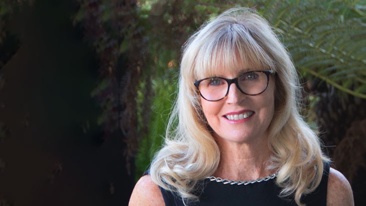 Smiling woman with long blonde hair and a fringe standing outdoors in front of greenery. She wears dark-framed glasses and a black top with a silver chain detail around the neckline.