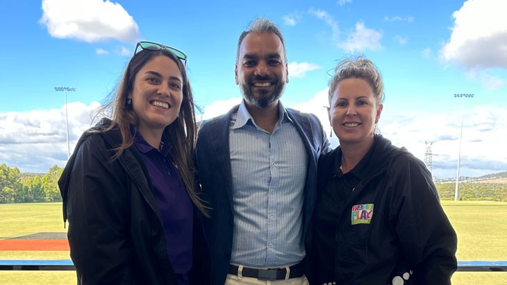 Three people are shown from the waist up standing outside. On the left is a woman with glasses on her head, in the middle a man wearing a button up shirt and blazer and on the right a woman with her hair up in a messy bun.