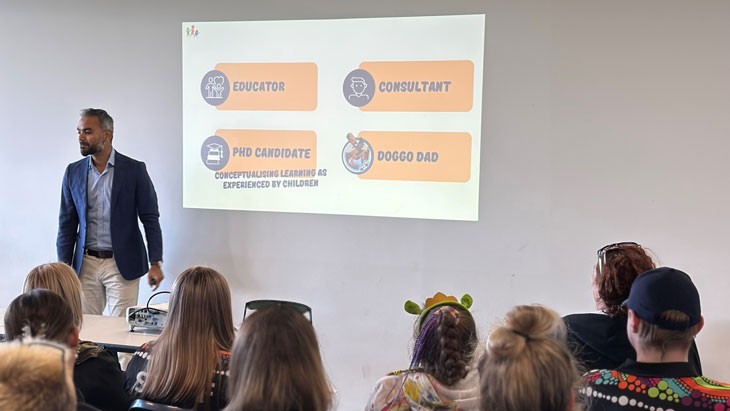 A group of people are seated looking at a PowerPoint presentation slide projected onto a light-coloured wall. A man with short black and white hair stands to the left of the presentation and addresses the group.