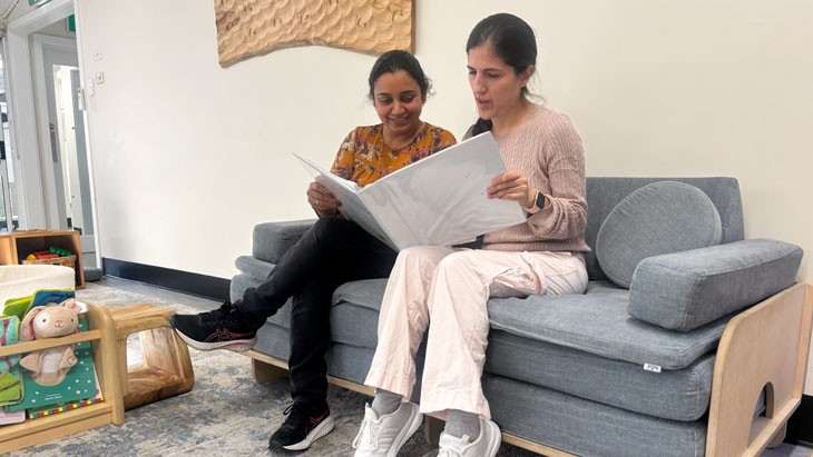 Two female educators sitting on a grey sofa reviewing policies and procedures in a white folder.