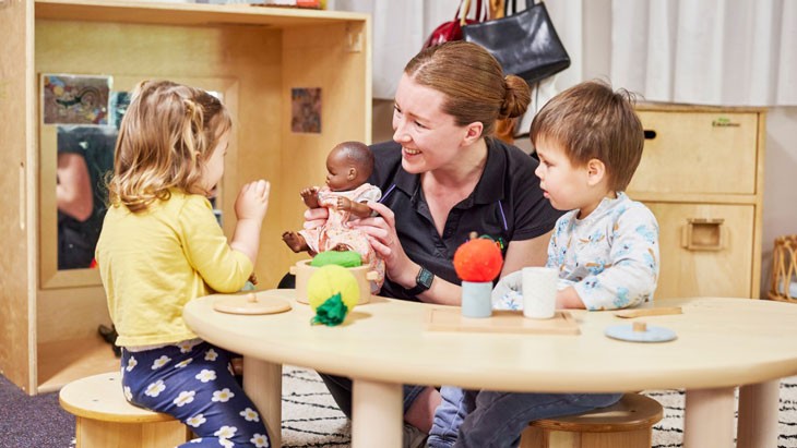 An educator and 2 children sit at a table playing with a doll and toy fruit.