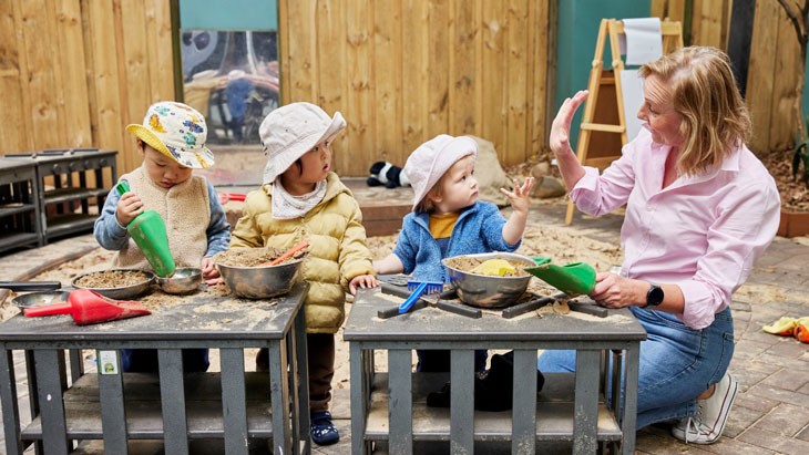 Three small children are engaged in sensory play outdoors, with an educator crouching beside them. Mixing bowls filled with sand and colourful scoops sit on the table in front of the children.