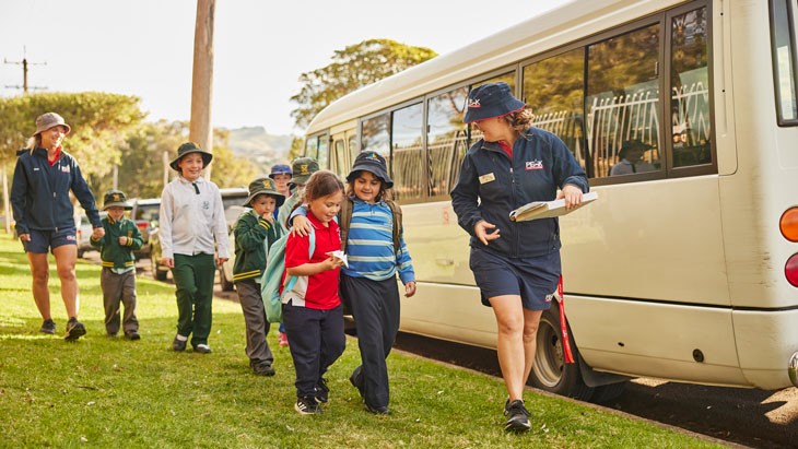 Young male student with short dark brown hair and wearing a blue school uniform sits on a mini bus, looking into the camera. He is surrounded by other young students wearing blue uniforms who are smiling and laughing.