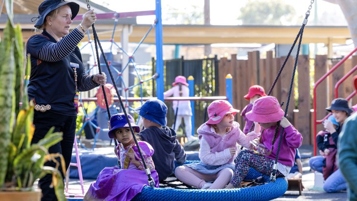 An educator stands next to children sitting on an accessible swing.