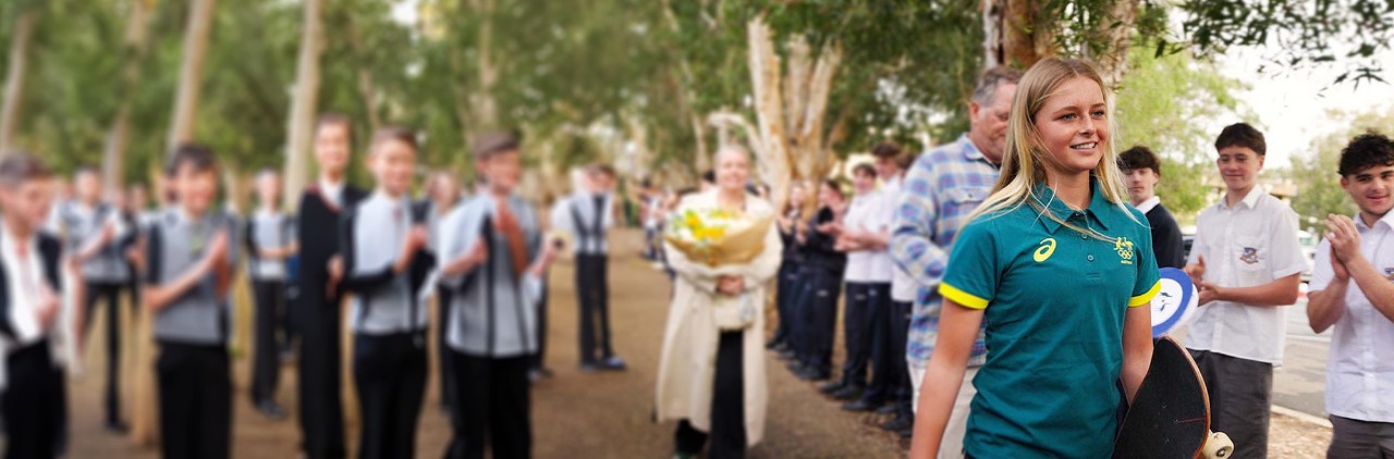  smiling blonde woman wearing a green Australian Olympic team polo shirt stands in the foreground of an outdoor scene, holding a bouquet of flowers while a diverse group of people—including school-uniformed students, men in suits and casual attire, and a woman in a long coat—applauds and stands behind her on a tree-lined path. The background features blurred green trees and a dirt ground, suggesting a school or community award ceremony.