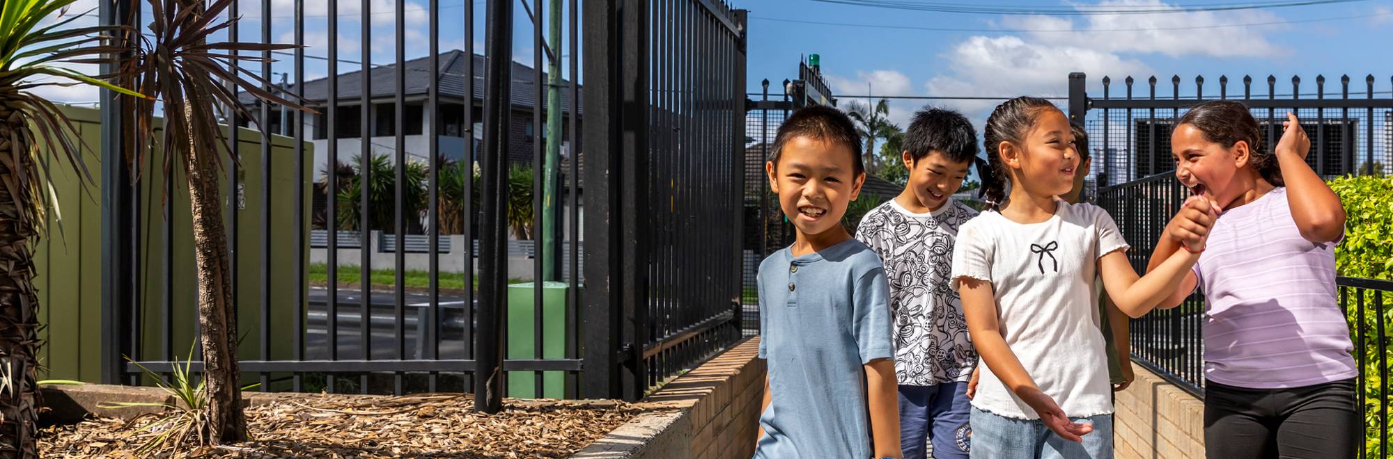 Three children walking and laughing along a school pathway beside a black fence in bright sunlight.