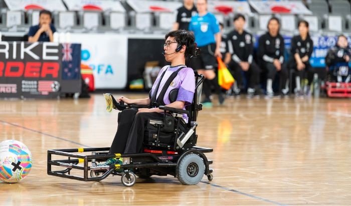 A young man wearing glasses in a wheelchair dressed in sports uniform playing powerchair football in an indoor arena