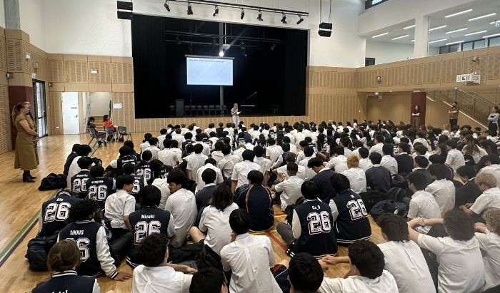 A big group of high school students with their backs to camera sitting on the floor of a school auditorium and listening to a woman speaking