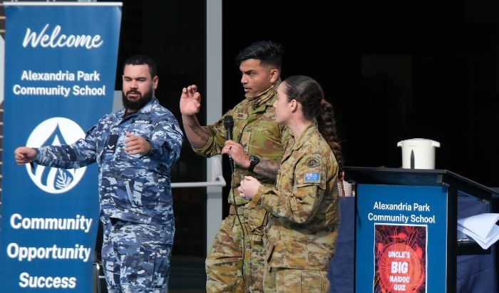 A woman and a man dressed in army khaki camouflage uniforms and another man dressed in army blue camouflage uniform presenting on a stage
