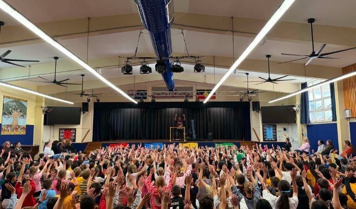 A big group of primary school students all with their arms raised in the air as they watch a performance of Aboriginal dancing on a stage in a school hall